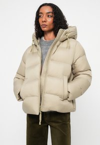 Chaqueta acolchada beige con capucha, que cuenta con un cierre frontal, bolsillos con botones y puños acanalados. Textura suave y acolchada.