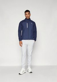 Tamsiai mėlynas vėjo nepraleidžiantis striukė su aukštu apykakliu ir priekyje užtrauktuku, derinamas su šviesiai pilkomis kelnėmis ir baltais sportbačiais. Matomi atspindintys akcentai.