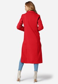 Manteau long en mélange de laine rouge avec un col cranté et une finition texturée, porté sur un jean bleu clair et des bottines crème.