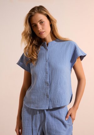 Jonge vrouw met golvend haar die een loszittend blauw textuur top met korte mouwen en knopen draagt, en bijpassende broek met trekkoord.
