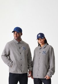 Δύο μοντέλα φορούν ασορτί καρό σακάκια με λογότυπα των New York Yankees και σκούρα μπλε καπέλα του baseball των Yankees, μπροστά από έναν απλό λευκό φόντο.