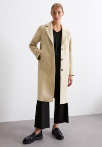 Knielanger, beiger Wollmantel mit Reverskragen, Fronttaschen und drei Knöpfen. Getragen über einem schwarzen Oberteil und weiten schwarzen Hosen.