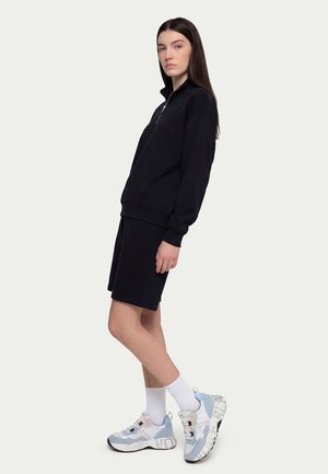 Kobieta stojąca w czarnej bluzie na zamek i spódnicy, ubrana w białe skarpetki i masywne białe i niebieskie buty sportowe, na prostym jasnym tle.