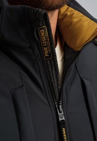 Chaqueta negra con forro amarillo, con un alto cuello, cierre de cremallera y tejido texturizado. La etiqueta dice "PME Legend American Classic."
