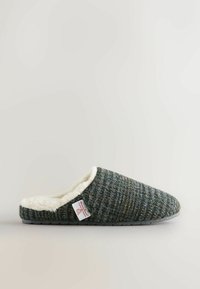 Slip-on hjemmesko med mørkegrønternet stofudvendig, hvid fluffy indvendig foring og en lille mærketag på siden.