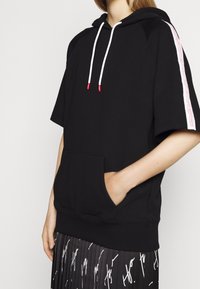 Sort hættetrøje med korte ærmer, der har en frontlomme, hvide snore med røde ender og et pink logo langs skuldersømmen.