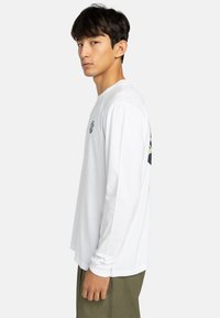 T-shirt in cotone bianco a maniche lunghe con una grafica sul retro, polsini a costine e scollo rotondo, indossato con pantaloni verde oliva.