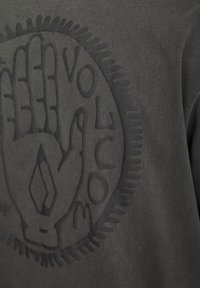 Felpa grigia scura con una grafica nera testurizzata di una mano e la parola "VOLCOM" in un design circolare. Materiale morbido in misto cotone.