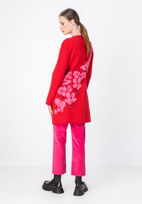 Červená pletená tunika s kvetinovým ružovým vzorom, skombinovaná s ružovými kordovým nohavicami a čiernymi robustnými topánkami. Hladká textúra, ležérny strih.