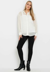 Durchsichtige weiße Bluse mit Knopfleiste, Chevron-Muster und Brusttasche, kombiniert mit schwarzen engen Jeans und schwarzen Knöchelstiefeln.