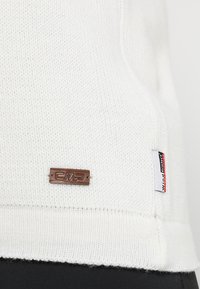 Ourlet de vêtement tricoté blanc avec logo CMP en métal et étiquette en tissu portant la mention « wind protect », porté sur un pantalon noir.