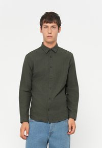 Giovane uomo che indossa una camicia verde scuro con bottoni e jeans azzurri chiari, in piedi di fronte a uno sfondo bianco semplice.