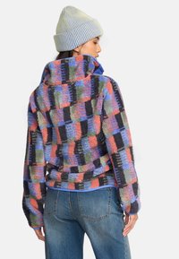Fleece pullover i et ternet mønster af pink, grøn, blå og sort, med høj krave og lange ærmer, parret med blå jeans.