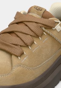 Καφέ σουέτ αθλητικό παπούτσι με πλεκτά καφέ κορδόνια, ραμμένες λεπτομέρειες και παχιά σκούρα σόλα. Λογότυπο στη γλώσσα και υφασμάτινη υφή κοντά στη φτέρνα.