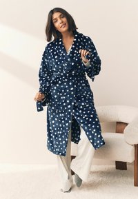 Tamsiai mėlynas minkštas chalatas su baltais taškeliais, su šaliku apykakle, raišteliu juosmeniui ir dviem priekinėmis kišenėmis. Modelis dėvi šviesiai pilkus kelnes.