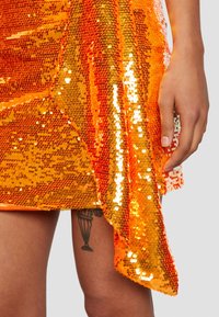 Vestido de lentejuelas naranja con un dobladillo asimétrico drapeado. Presenta lentejuelas brillantes y reflectantes, además de una textura suave. Mano apoyada sobre la tela.