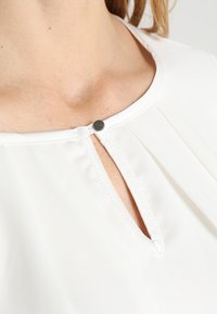 Vit blus med nyckelhålsutskärning, med en liten silverknapp, veckade detaljer vid kragen och slät tygtextur.