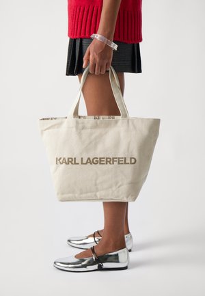Borsa tote in tela beige con "KARL LAGERFELD" in grassetto marrone. Presenta due manici corti e un interno a fantasia. Indossate ballerine argento.