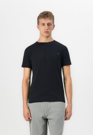 Young man with short hair wearing a plain black short-sleeve t-shirt and light grey pants, standing against a white background.