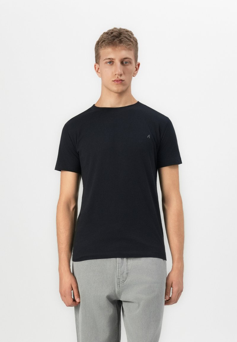 Tânăr cu părul scurt, îmbrăcat cu un tricou simplu negru cu mânecă scurtă și pantaloni gri deschis, stând pe un fundal alb.