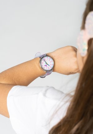 Montre pour enfants avec un cadran rond rose et blanc, de grands chiffres noirs, un bracelet en plastique transparent et des accents décoratifs en forme de cœur.