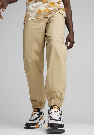 Beige joggerbukser har elastiske mansjetter og sidelommer. Båret med sorte, hvide og orange sneakers. Baggrunden er neutral grå.
