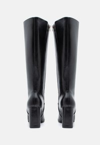 Μαύρες δερμάτινες μπότες μέχρι το γόνατο με λεία υφή, τετράγωνα τακούνια και φερμουάρ στο πίσω μέρος. Ο κορμός έχει σχεδίαση με γωνιώδη κορυφή.