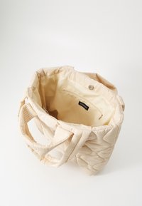 Sac cabas matelassé beige avec un extérieur texturé, deux poignées et un intérieur spacieux comprenant une poche zippée et une étiquette de marque.