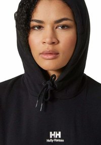 Czarna bluza z kapturem i ściągaczem. Posiada białe logo Helly Hansen na klatce piersiowej oraz miękką teksturę. Zbliżenie na twarz.