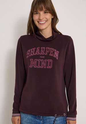 Femme souriante portant un t-shirt à manches longues violet foncé avec le texte "Sharpen Your Mind" et un jean bleu sur un fond uni.
