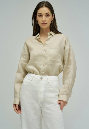 Mujer con camisa beige de manga larga abotonada, metida dentro de pantalones blancos de cintura alta, de pie contra un fondo claro y liso.