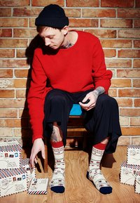 Czerwony sweter o teksturowanej powierzchni, czarna czapka beanie, luźne czarne spodnie, wzorzyste kremowe skarpetki z czerwonymi i granatowymi akcentami, otoczony pudełkami prezentowymi na drewnianej podłodze.