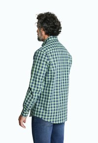 Hombre con cabello rizado y barba, viste una camisa de cuadros verde y azul y jeans azules, de espaldas contra un fondo blanco.