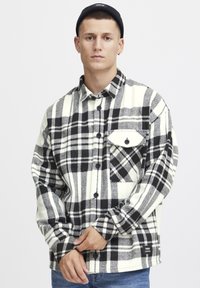 Camicia flanella a quadri bianco e nero con maniche lunghe, chiusura con bottoni, taschino sul petto e texture morbida. Indossata sopra jeans blu.