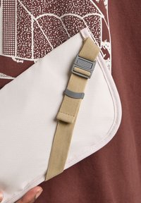 Witte textuur zak met beige riem en grijze hardware, met een afgeronde rand en zichtbare stiksels, gehouden tegen een roodachtige achtergrond.