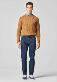 Bruine kabelgebreide trui, blauwe overhemden, donkere spijkerbroek en sneakers. De trui heeft een gestructureerd patroon; de outfit is casual met een getailleerde look.