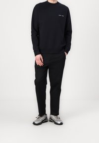 Hombre vistiendo un sudadera negra con un pequeño texto blanco, pantalones negros y zapatillas deportivas plateadas, de pie contra un fondo blanco liso.