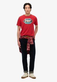 Camiseta de algodón roja con un logo gráfico, combinada con unos pantalones vaqueros oscuros y una camisa de cuadros roja y negra atada a la cintura. Zapatillas blancas.