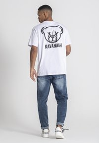 Camiseta blanca de algodón con un gran gráfico de un oso y el texto "KAVANAGH" en la parte posterior. Combinada con unos vaqueros azules y zapatillas blancas.