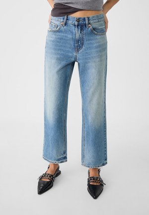 Vrouw die lichtblauwe jeans met rechte pijpen en zwarte platte schoenen met puntige neus en met studs versierde riemen draagt, staand tegen een grijze achtergrond.
