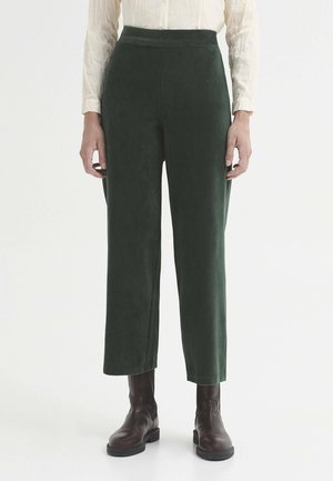 Femme portant un pantalon court en velours côtelé vert foncé taille haute et des bottines marron foncé, debout devant un fond uni.