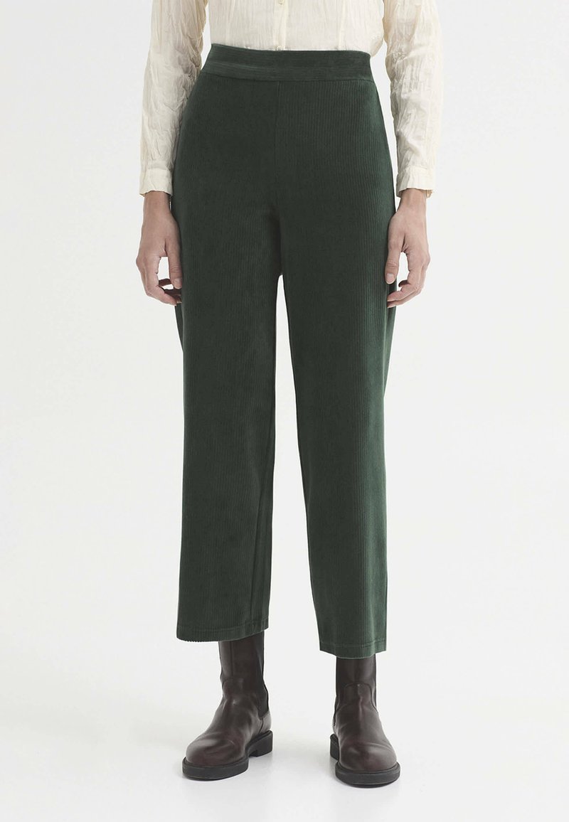 Femme portant un pantalon court en velours côtelé vert foncé taille haute et des bottines marron foncé, debout devant un fond uni.