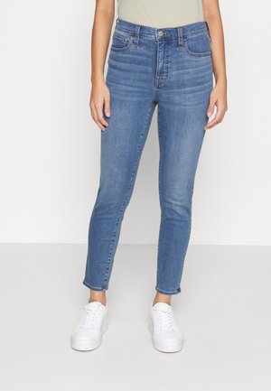 Blå denimjeans med hög midja, slim fit och ankellängd, tillsammans med vita sneakers och en ljusgrön topp.