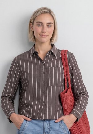 Kvinde med kort blondt hår iført en brun og hvidstribet skjorte, blå jeans og bærende en rød strikket skuldertaske.