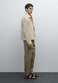 Beige lněný sako, khaki košile, hnědé kalhoty a černé sandály. Střih je volný, s knoflíkovými manžetami a detaily na popruzích sandálů.