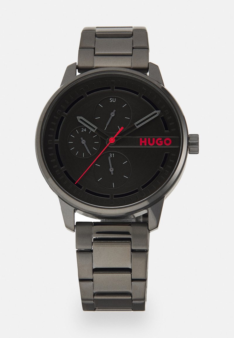 Reloj de metal negro con una esfera redonda, tres subesferas y un logotipo HUGO rojo. Presenta una correa de tono plateado y acentos rojos contrastantes.