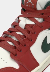 Crvene i bijele visoke tenisice s crvenim vezicama i zelenim Nike swoosh detaljem, prikazane iz blizine, fokus na vezivanje i gornju stranu.