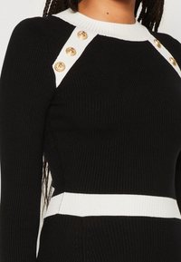 Vestido negro acanalado con acentos blancos en los hombros y la cintura, que presenta cuatro botones dorados a lo largo de las costuras de los hombros.