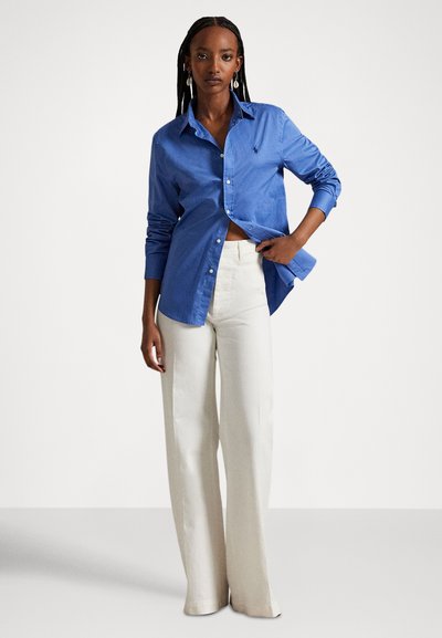 Nő hosszú fonatokkal, kék gombos ingben és magasderekú, bő szárú fehér nadrágban áll, sima fehér háttér előtt.