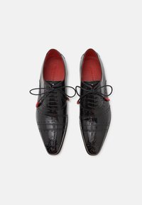 Scarpe da uomo in vernice nera con punte appuntite, dettagli in brogue e fodera interna rossa. Presentano lacci neri e dettagli di accentuazione.